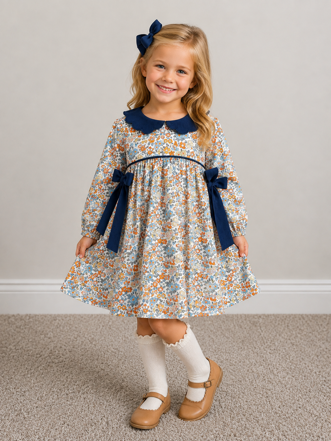A young girl with blonde hair wearing a long-sleeve floral dress with a navy blue collar and bows, white knee-high socks, and tan Mary Jane shoes, standing on a carpeted floor against a plain wall.