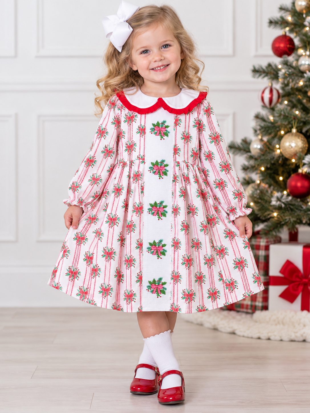 A young girl wearing a festive holiday dress with a red and white striped pattern, green holly motifs, and a red collar. She is standing in a decorated room next to a Christmas tree.
