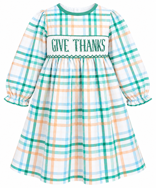 A long-sleeve plaid dress with a white smocked chest panel that reads 'GIVE THANKS' in green lettering. The dress features a white base with green, light blue, and peach plaid lines, and a green collar.