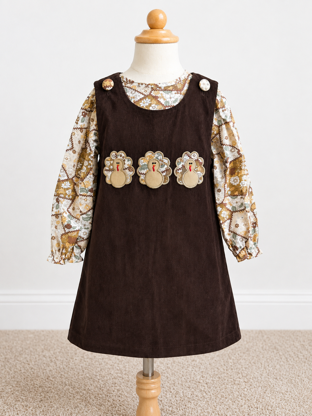 A toddler girl's dress displayed on a wooden dress form against a white wall and beige carpet. The dress has a dark brown corduroy pinafore-style front with three turkey appliques across the chest and a long-sleeve underlayer in a beige, brown, and gold patterned print.
