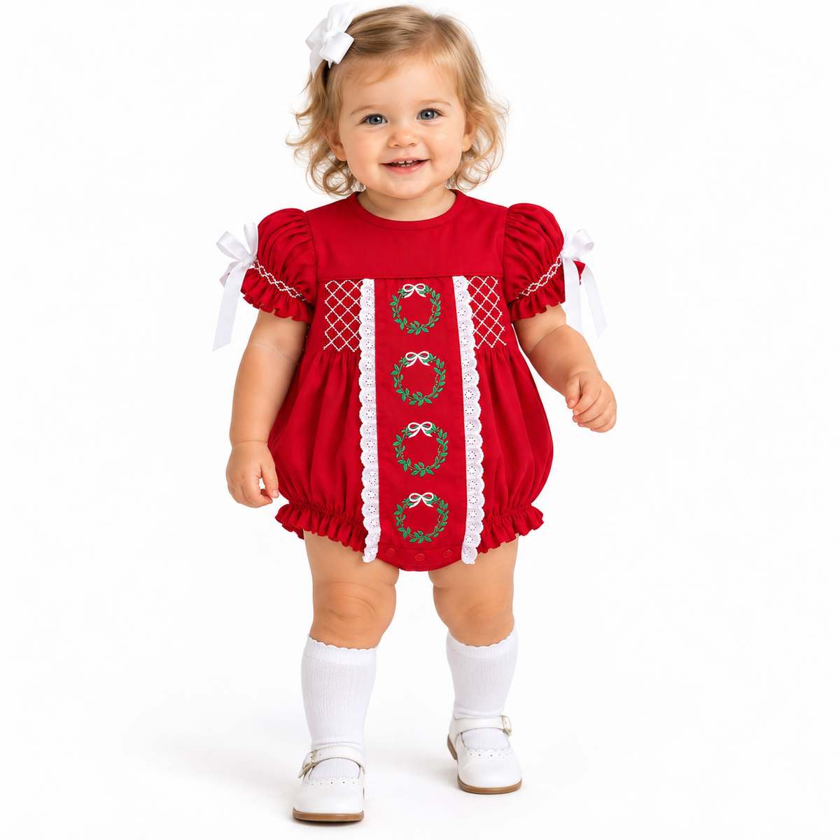 Toddler girl wearing a red Christmas bubble with embroidered green wreaths, white lace trim and white ribbon sleeve ties.