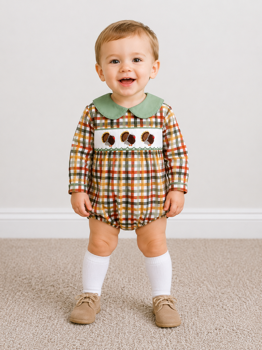 A toddler standing on a beige carpet against a white wall, wearing a long-sleeve plaid romper with a green Peter Pan collar and an embroidered chest panel with acorns. The romper has a multicolor plaid pattern in orange, green, yellow, and brown.