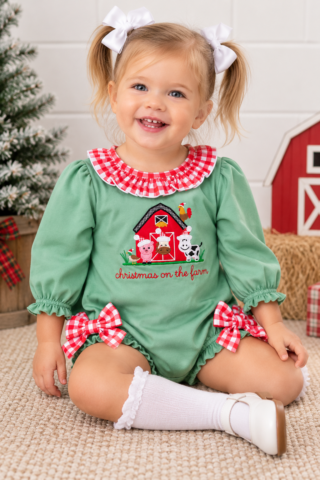A toddler girl with blonde pigtails sits on a beige carpet wearing a green long-sleeve romper with red and white gingham trim, a red barn and farm animal applique on the chest, and red gingham bow accents on the legs. She is wearing white knee-high socks and white shoes, with a Christmas tree and barn decorations in the background.