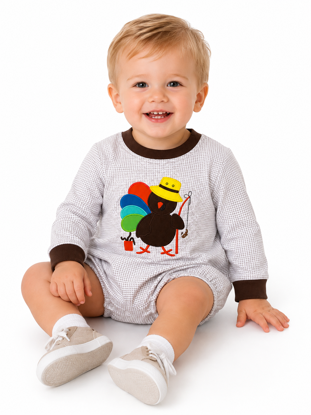 A smiling baby boy wearing a white and gray checkered long-sleeve romper with brown trim and a turkey graphic, sitting against a white background.